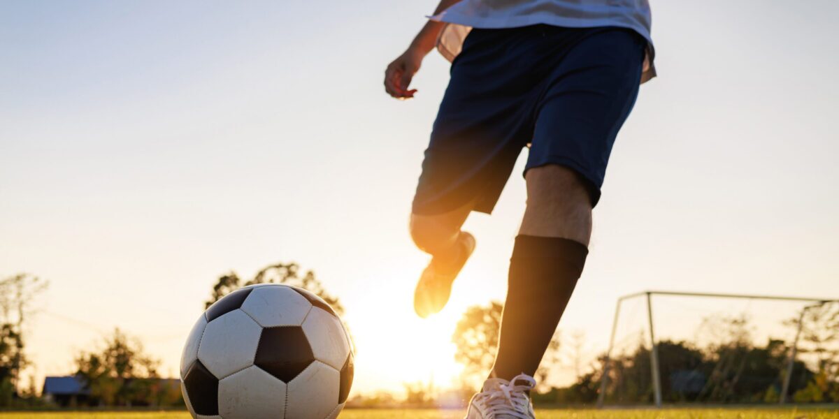 Close up shot of soccer player kicking ball. Action sport outdoors playing football for exercise at green grass field under the twilight sunset.