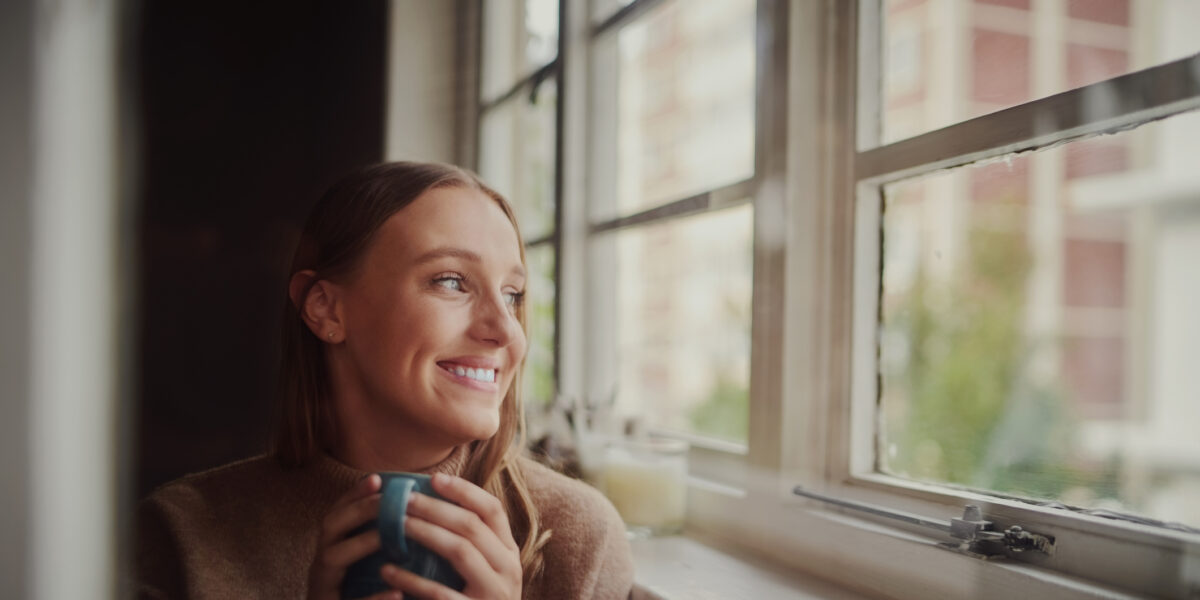 Quarantined young woman smiling drinking coffee and daydreaming looking out at empty streets