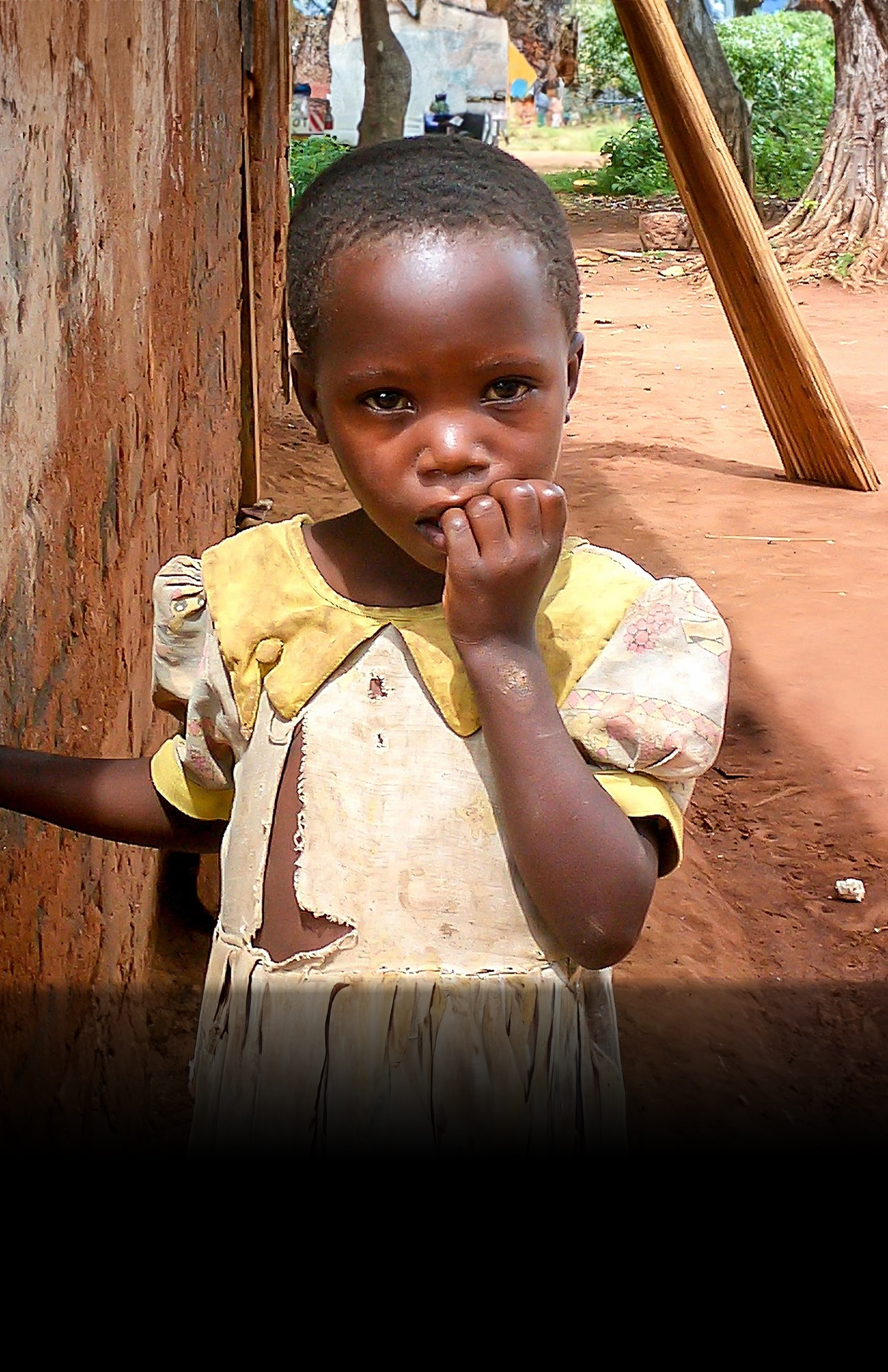 Young child in yellow dress standing by wooden post with hand to mouth in rural setting