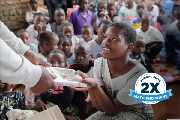 Child seated among other children, receiving a book from an adult.