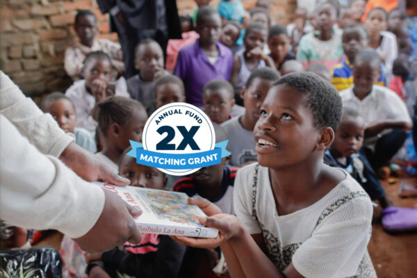 Child seated among other children, receiving a book from an adult.