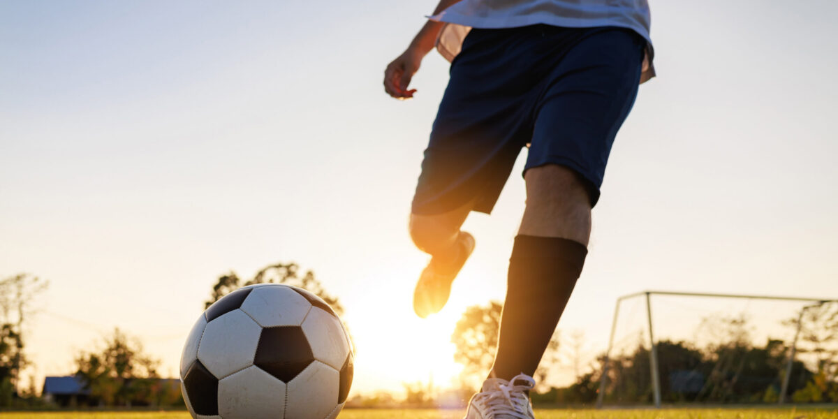 Close up shot of soccer player kicking ball. Action sport outdoors playing football for exercise at green grass field under the twilight sunset.